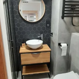 Nowoczesna łazienka z czarnymi heksagonalnymi płytkami, okrągłym lustrem, drewnianą szafką pod umywalką i czarnym grzejnikiem drabinkowym.