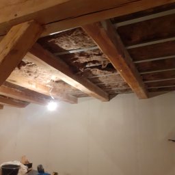 Mar-Bud - Drewniane belki stropowe z izolacją z wełny mineralnej, widoczne elementy metalowej konstrukcji podtrzymującej, na podłodze narzędzia i materiały budowlane, ściany przygotowane do dalszych prac...