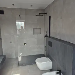 Nowoczesna łazienka wykończona w szarościach z czarnymi akcentami: deszczownica, bateria umywalkowa i bidetowa, toaleta podwieszana. W lustrze odbija się osoba robiąca zdjęcie.