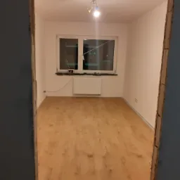 Pokój po ułożeniu paneli podłogowych, widok zza ościeżnicy, z grzejnikiem pod oknem i pojedynczą lampą sufitową.