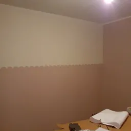 Pomieszczenie po częściowym malowaniu ścian na dwa kolory, z falistym wzorem oddzielającym barwy, widoczne materiały malarskie na podłodze.