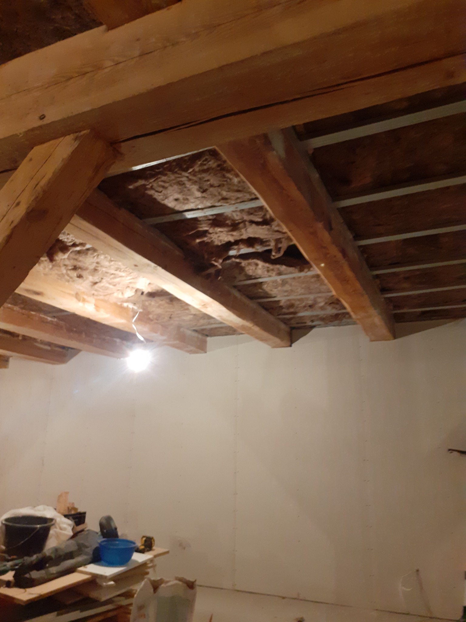 Drewniane belki stropowe z izolacją z wełny mineralnej, widoczne elementy metalowej konstrukcji podtrzymującej, na podłodze narzędzia i materiały budowlane, ściany przygotowane do dalszych prac...