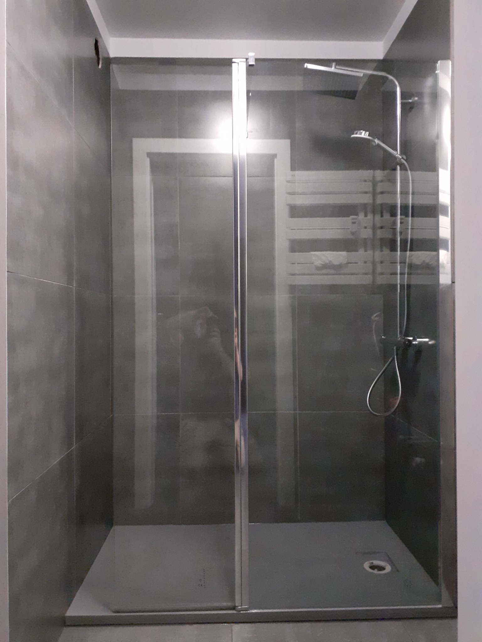 Nowoczesna kabina prysznicowa z szarymi płytkami, szklanymi drzwiami i chromowaną armaturą, widok z przodu na brodzik i odpływ.