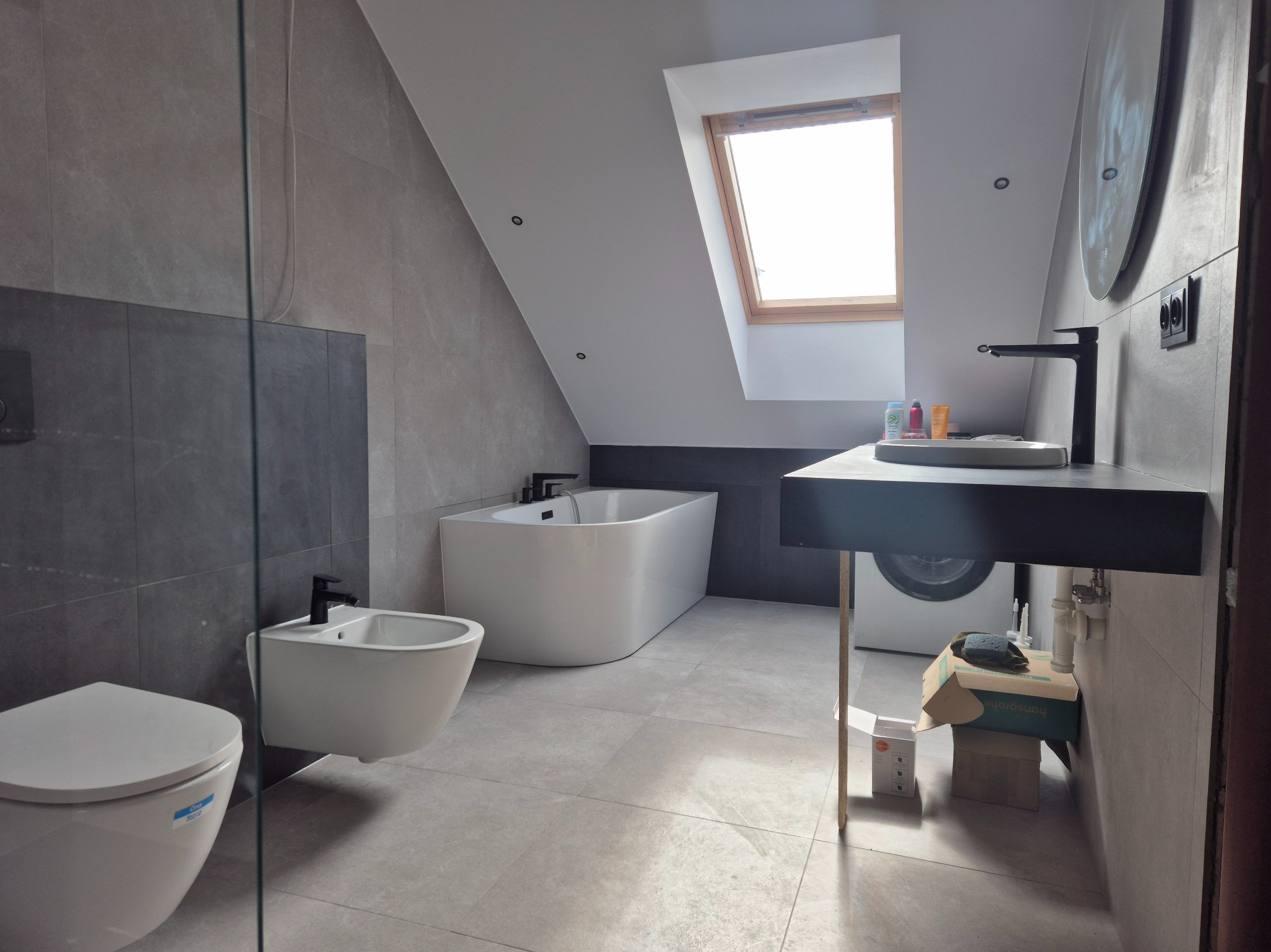 Nowoczesna łazienka na poddaszu z oknem dachowym, białą wanną wolnostojącą, wiszącą toaletą i bidetem, umywalką nablatową na czarnym blacie oraz szarą płytką ceramiczną na ścianach i podłodze.