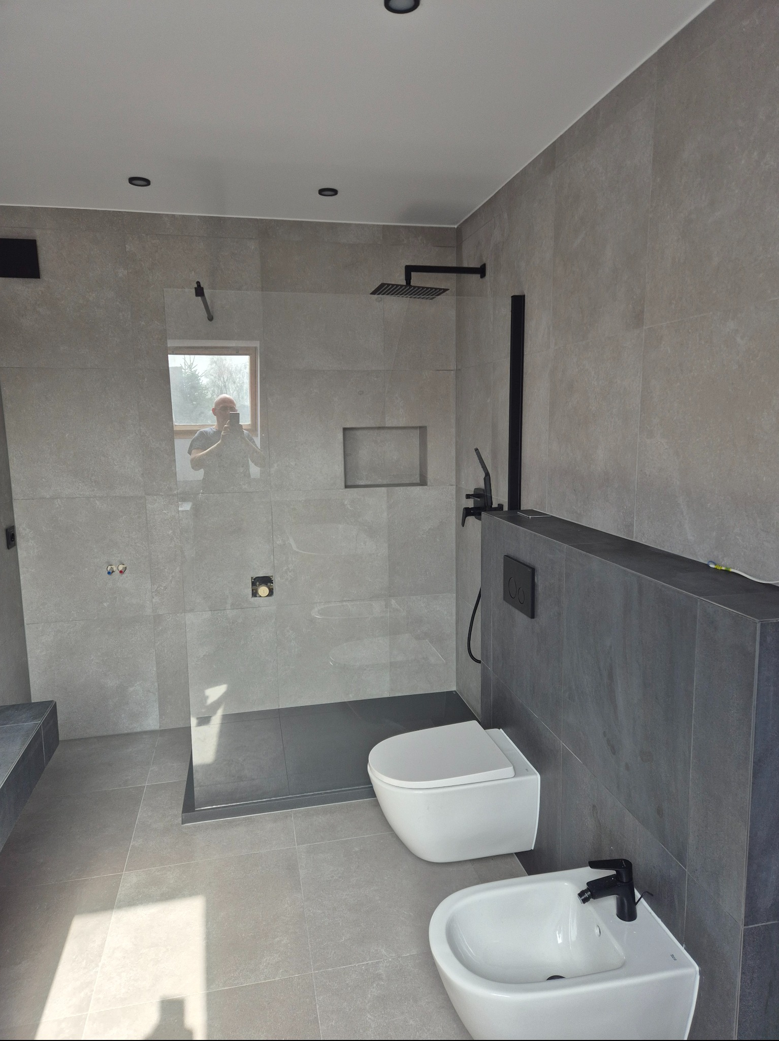 Nowoczesna łazienka wykończona w szarościach z czarnymi akcentami: deszczownica, bateria umywalkowa i bidetowa, toaleta podwieszana. W lustrze odbija się osoba robiąca zdjęcie.