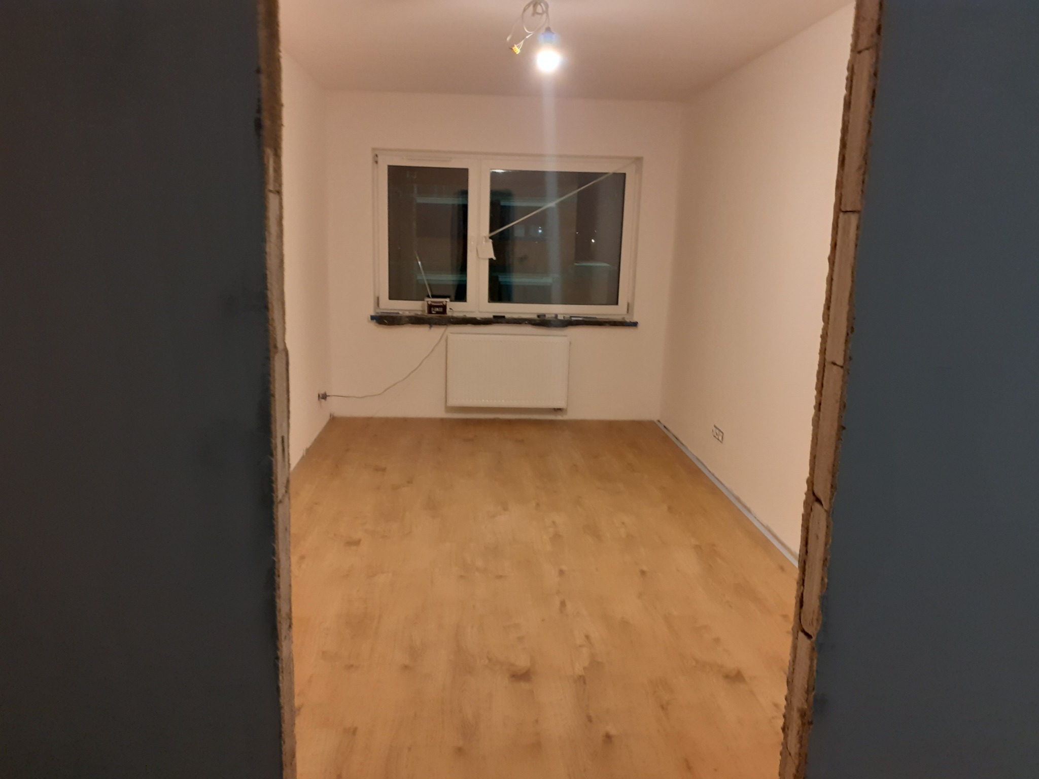 Pokój po ułożeniu paneli podłogowych, widok zza ościeżnicy, z grzejnikiem pod oknem i pojedynczą lampą sufitową.