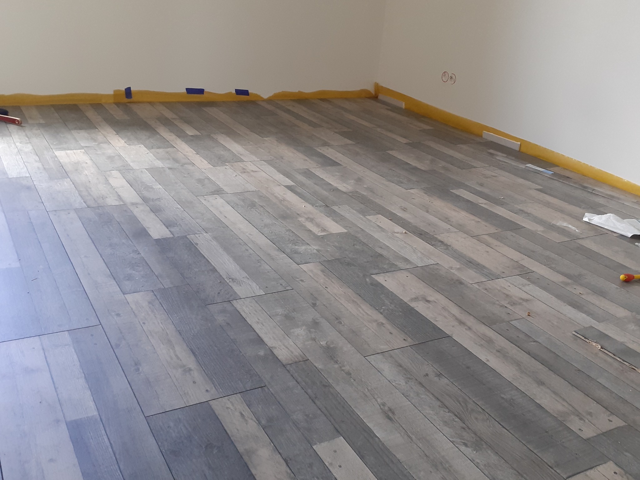 Ułożone panele podłogowe w szaro-brązowej tonacji, zabezpieczone taśmą malarską przy ścianach, widoczne gniazdka elektryczne i drobne narzędzia.