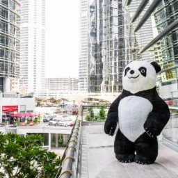Duży dmuchany kostium pandy na tle nowoczesnej architektury miejskiej, widoczny z tarasu z metalową barierką.