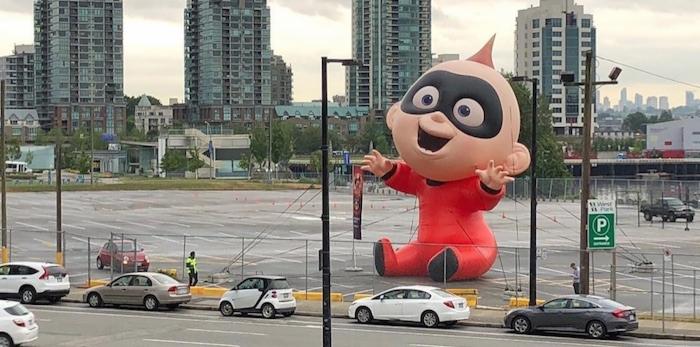 Ogromna dmuchana postać Jack-Jack z filmu Iniemamocni siedzi na parkingu, w tle miejski krajobraz z wieżowcami i pochmurne niebo.