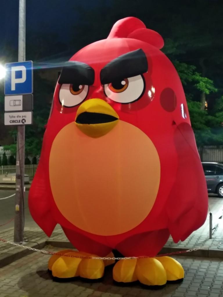 Duży, czerwony dmuchany ptak z gry Angry Birds stoi na chodniku przy znaku parkingu Circle K, otoczony łańcuchem.