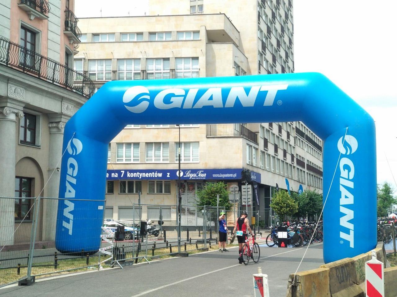 Niebieski dmuchany łuk z logo Giant ustawiony na ulicy podczas wydarzenia rowerowego, widoczni rowerzyści i zaparkowane rowery.
