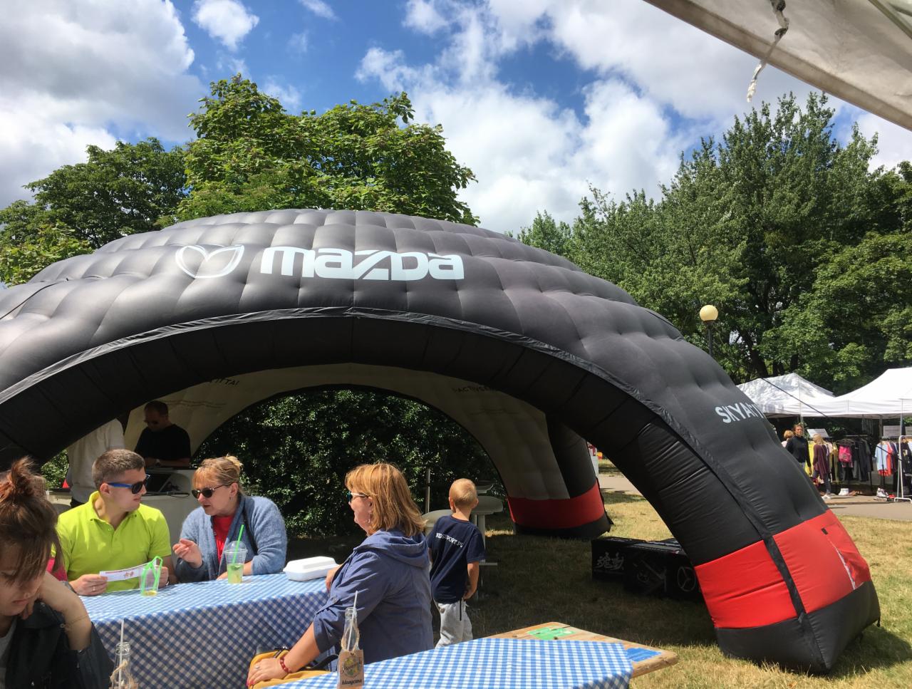Czarny dmuchany namiot z logo Mazda na imprezie plenerowej, osoby siedzące przy stolikach z niebiesko-białymi obrusami, tło z zielenią drzew i błękitnym niebem z chmurami.
