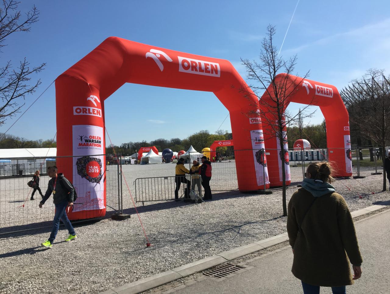 Dwa czerwone dmuchane łuki z logo Orlen Warsaw Marathon na terenie imprezy plenerowej z widocznymi osobami i namiotami w tle.