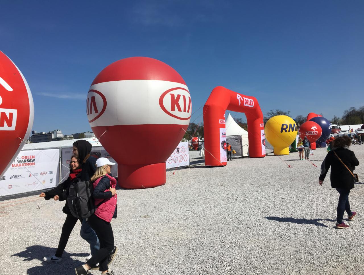 Uczestnicy Orlen Warsaw Marathon przechodzą obok dmuchanych bramek i balonów reklamowych marek Kia i Orlen na żwirowym terenie.