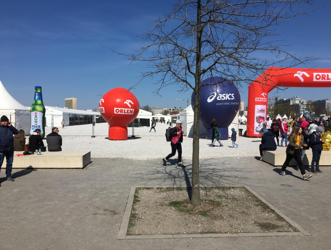 Uczestnicy biegu miejskiego przechodzą obok dmuchanych reklam Orlen i Asics, widoczny również dmuchany model butelki piwa Lech.