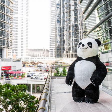 Duży dmuchany kostium pandy na tle nowoczesnej architektury miejskiej, widoczny z tarasu z metalową barierką.