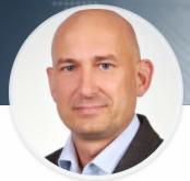 Portret mężczyzny z łysą głową, ubranego w ciemną marynarkę, na białym tle w okrągłej ramce. Zdjęcie profilowe.