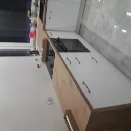 Nowoczesna zabudowa kuchenna z białymi frontami i drewnopodobnym blatem, widoczne gniazdka elektryczne na ścianie oraz okno z widokiem na zewnątrz.