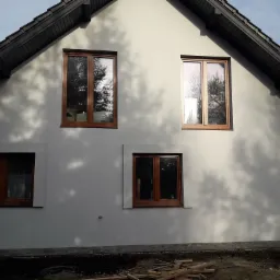 Elewacja domu jednorodzinnego w jasnym odcieniu szarości z drewnianymi oknami, czarnym dachem i widocznymi refleksami drzew na ścianie, z fragmentem zaparkowanego białego samochodu dostawczego po...