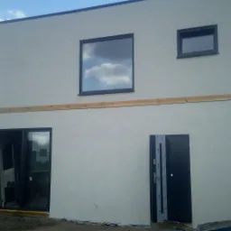 Surowa elewacja domu z zamontowanymi oknami i drzwiami, widoczne drewniane elementy konstrukcyjne przed wykończeniem, teren wokół domu w trakcie prac ziemnych.