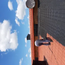 Pracownik w trakcie montażu ciemnoszarej blachodachówki na czerwonym dachu, widoczne narzędzia i fragment muru z cegły na tle błękitnego nieba z chmurami.