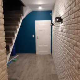 Korytarz po remoncie z białą cegłą na ścianie, drewnianymi schodami i niebieską ścianą w tle, z nowoczesnym oświetleniem.