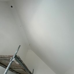 DOMIZ DOMINIK KINEL - Świeżo pomalowane, białe poddasze ze skosami, widoczne ślady po szpachlowaniu i punktowe oświetlenie, częściowo zasłonięte przez metalowe rusztowanie.