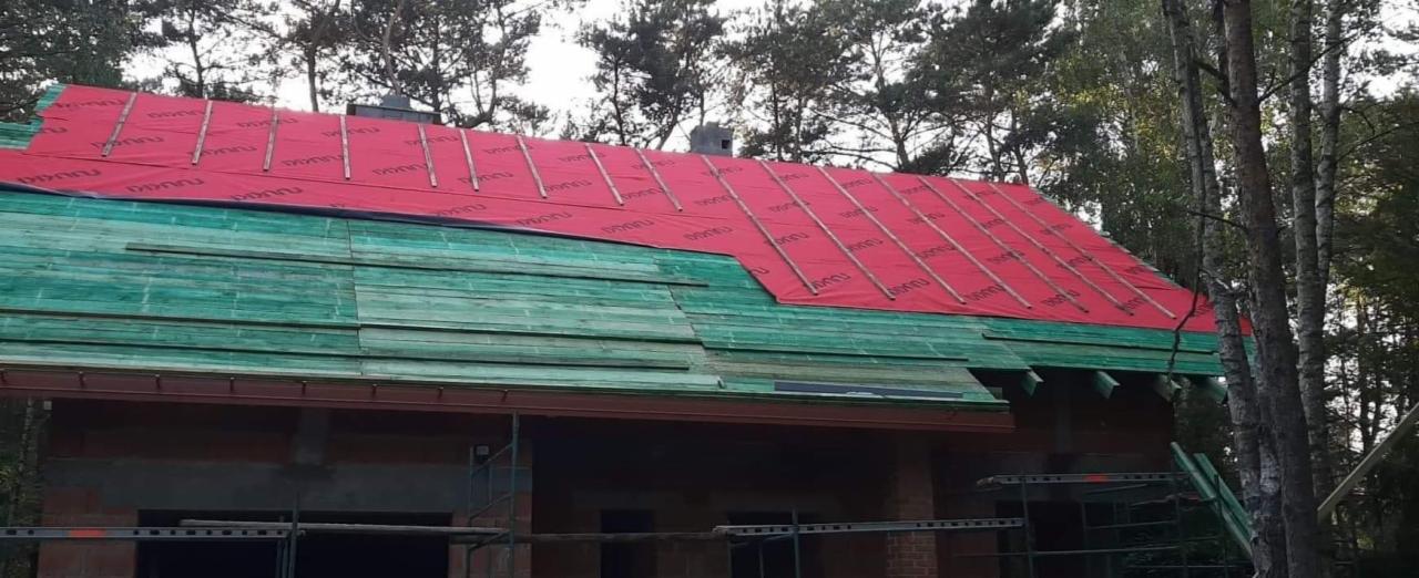 Budowa dachu: widok na drewniany dach z częściowo ułożoną czerwoną membraną i listwami kontrłat, dom w stanie surowym otoczony drzewami.