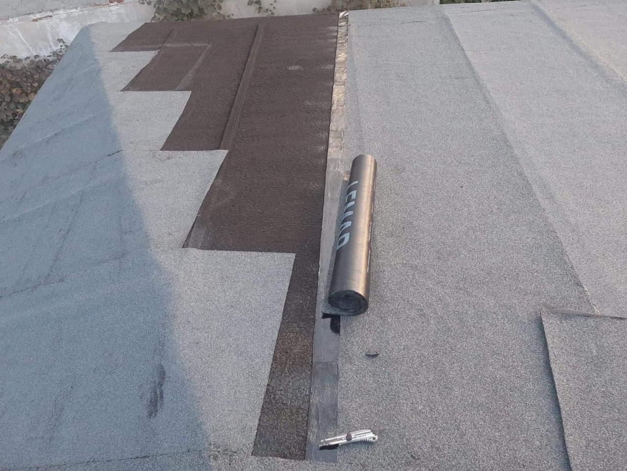 Fragment płaskiego dachu pokrytego papą, widoczny częściowo nowy pas papy ułożony schodkowo, obok rolka papy i nóż monterski.