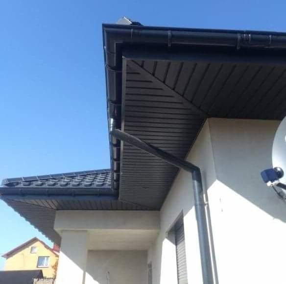 Fragment domu jednorodzinnego z czarną, metalową rynną, podbitką i dachem krytym dachówką ceramiczną w odcieniach szarości, widziany w słoneczny dzień.