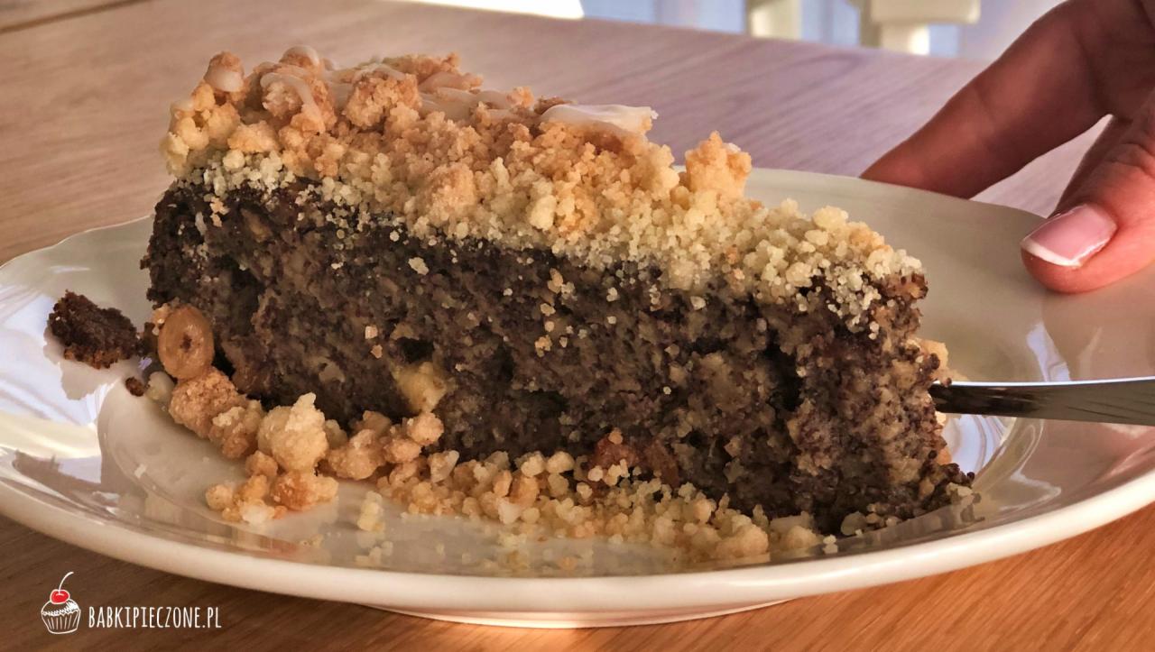 Kawałek makowca z kruszonką i lukrem, podawany na białym talerzu, z widoczną ręką osoby trzymającej widelec.