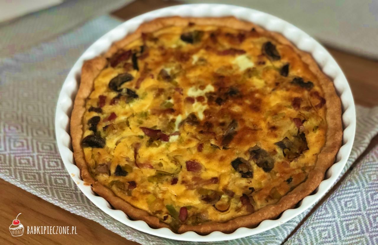 Quiche Lorraine z boczkiem, pieczarkami i porem w białej formie na wzorzystej ściereczce.