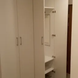 Biała szafa wnękowa z otwartymi drzwiami, widoczne półki i skrzynka elektryczna wewnątrz, szare płytki podłogowe.