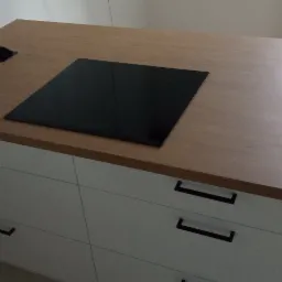 Blat drewniany z wbudowaną płytą indukcyjną i gniazdkami, umieszczony na białych szafkach z ciemnymi uchwytami.