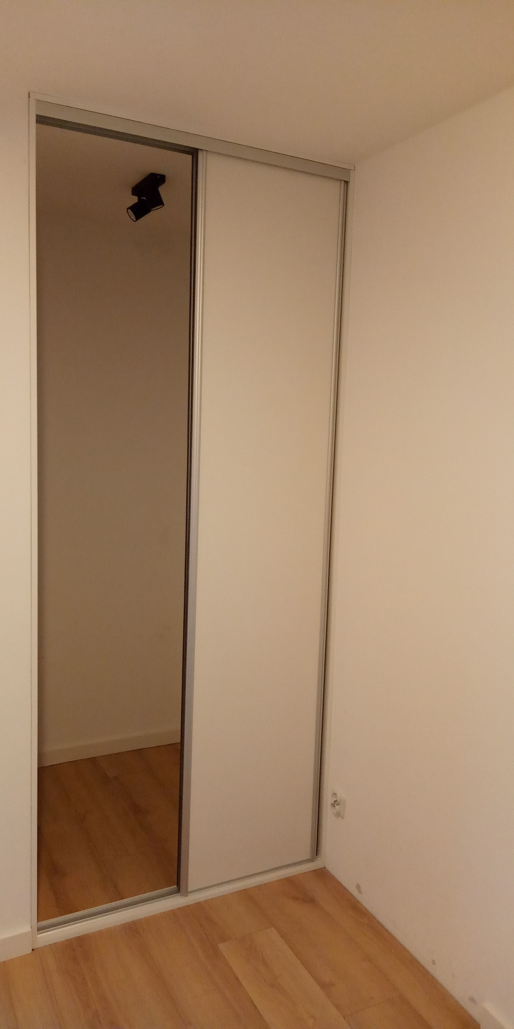 Biała szafa przesuwna z lustrzanymi drzwiami w rogu pokoju, widoczne oświetlenie punktowe na suficie wewnątrz szafy.