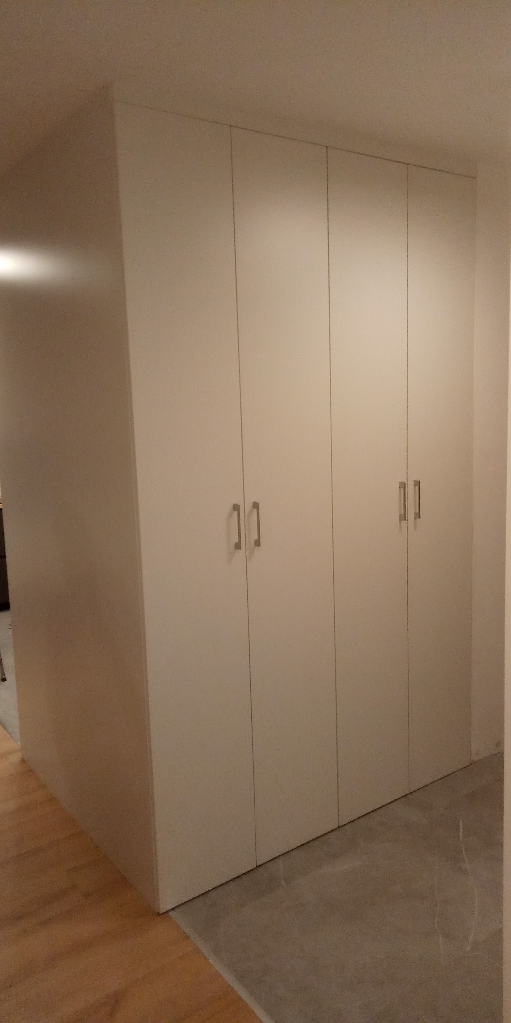 Biała szafa wnękowa z czterema drzwiami i minimalistycznymi uchwytami, dopasowana do sufitu, z widoczną podłogą z dwóch rodzajów materiałów.