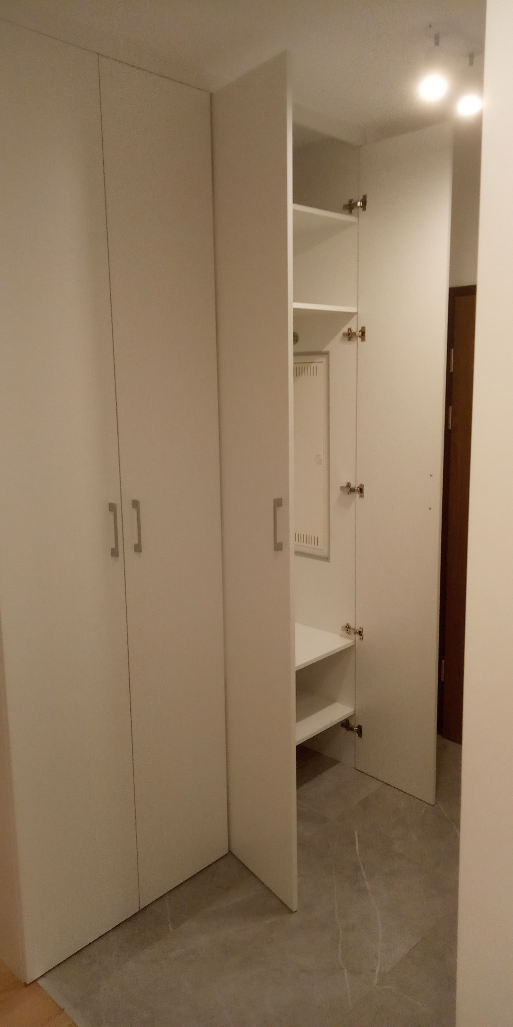 Biała szafa wnękowa z otwartymi drzwiami, widoczne półki i skrzynka elektryczna wewnątrz, szare płytki podłogowe.