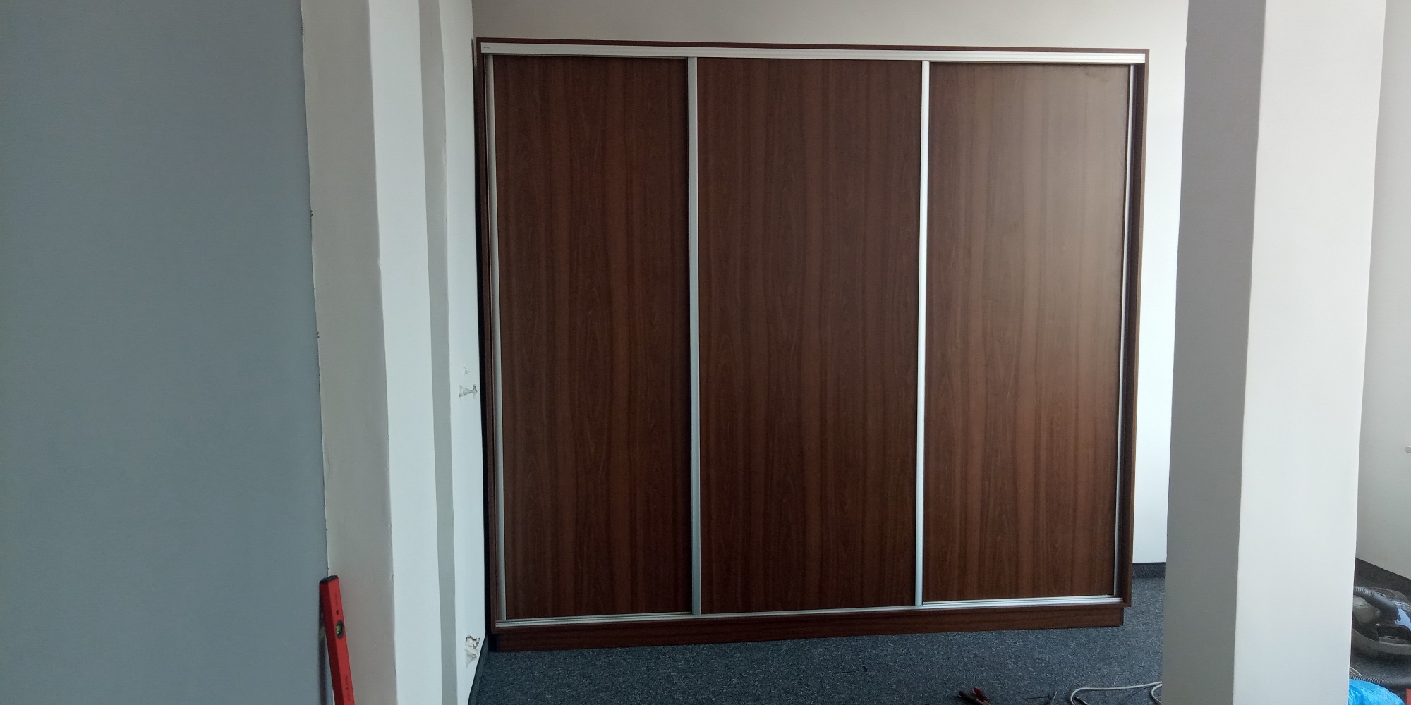 Trzydrzwiowa szafa przesuwna z okleiną drewnopodobną w odcieniach brązu, widoczna z perspektywy wnętrza, z poziomnicą opartą o ścianę po lewej stronie.