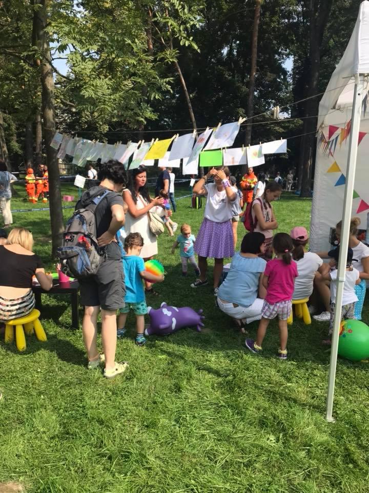 Plenerowa impreza z dziećmi na trawie; na sznurku wiszą rysunki dzieci; w tle drzewa i namiot.