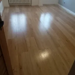 Jasne panele podłogowe ułożone w pustym pokoju z dwoma oknami i grzejnikiem, widoczne niedoskonałości łączeń paneli.