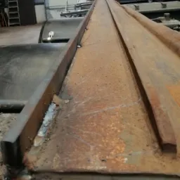 Fragment konstrukcji stalowej z widocznymi śladami spawania, pokrytej rdzą i zabrudzeniami, w tle drabina i elementy warsztatu.