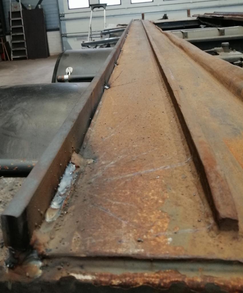 Fragment konstrukcji stalowej z widocznymi śladami spawania, pokrytej rdzą i zabrudzeniami, w tle drabina i elementy warsztatu.