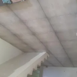 Widok na sufit z szarymi płytkami, z widocznymi sznurkami dystansowymi, fragmentem ściany i schodów w trakcie prac wykończeniowych.