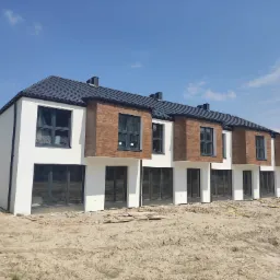 Nowoczesne segmenty w zabudowie szeregowej z białymi ścianami i drewnopodobną elewacją, czarną dachówką i dużymi oknami, teren wokół w trakcie prac wykończeniowych.