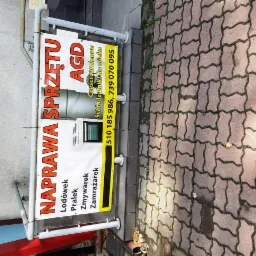 Baner reklamowy firmy oferującej naprawę sprzętu AGD, w tym lodówek, pralek, zmywarek i zamrażarek, z numerami telefonów kontaktowych, umieszczony przed budynkiem.