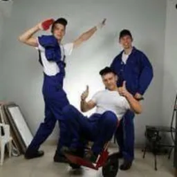 Trzech młodych mężczyzn w niebieskich kombinezonach pozuje w studiu: jeden stoi z ręką uniesioną nad głową trzymając czerwoną szmatkę, drugi siedzi na czerwonym w&oacute;zku z uniesionymi kciukami,...