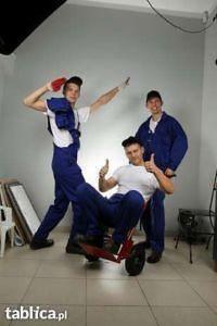 Trzech młodych mężczyzn w niebieskich kombinezonach pozuje w studiu: jeden stoi z ręką uniesioną nad głową trzymając czerwoną szmatkę, drugi siedzi na czerwonym wózku z uniesionymi kciukami,...