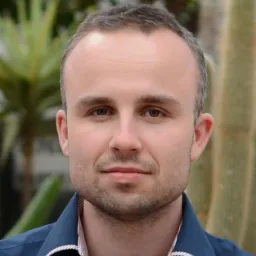 Portret mężczyzny w niebieskiej koszuli z roślinami w tle, lekki zarost, skupione spojrzenie.