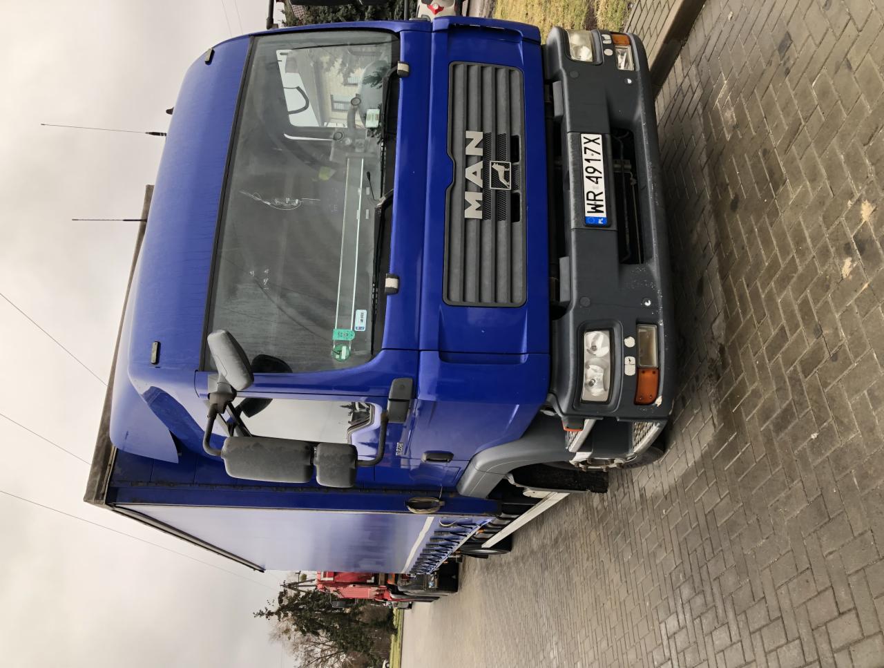 Niebieska ciężarówka MAN z plandeką, widok z przodu, tablica rejestracyjna WR 4917X, zaparkowana na brukowanej nawierzchni w pochmurny dzień.