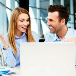 Uśmiechnięta kobieta z zegarkiem na ręce gestykuluje, rozmawiając z mężczyzną pochylonym nad laptopem w nowoczesnym biurze, dokumenty i telefon na stole.